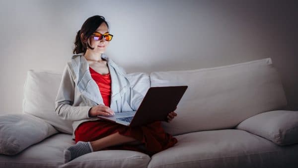 Young woman with blue light blocking glasses (yellow lenses) working with laptop on white sofa