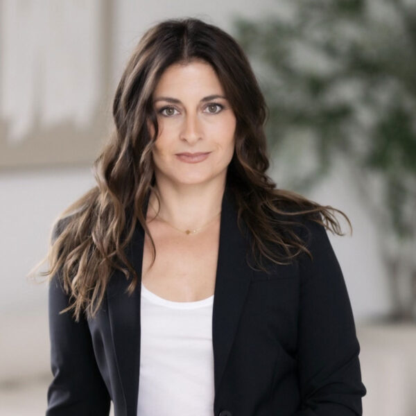 Portrait of a woman with long wavy brown hair, wearing a black blazer and white top, looking at the camera in a neutral setting.